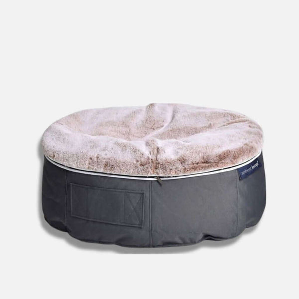 En rund Ambient Lounge-hundeseng med en blød, beige, fluffy top og en mørkegrå stofbund, med en lille sidelomme og etiket på siden, placeret på en lys baggrund.