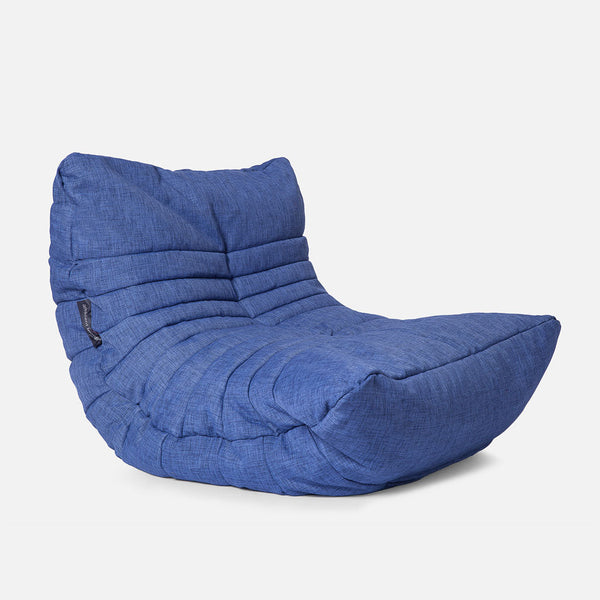 En moderne salongstol i blåt stof med lavt sæde, armløst design og blød, polstret tekstur. Stolen har vandrette syninger og en afslappet, uformel form, der minder om en saccosekk.