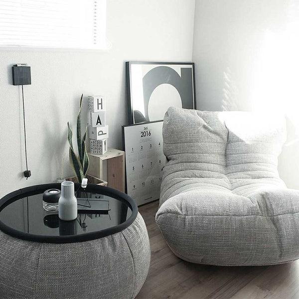 Et lyst, minimalistisk rum med en hyggelig grå salongstol, et rundt sort sofabord med en vase og et stearinlys, en slangeplante og indrammet kunst og en stor kalender, der står op ad væggen på trægulvet.