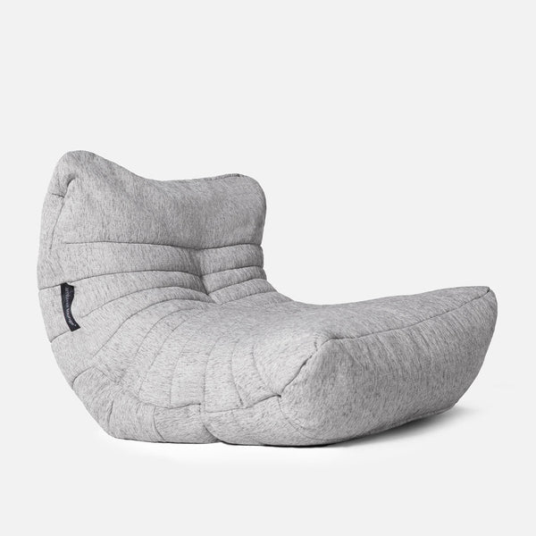 En moderne, lysegrå salongstol med lavt sæde, bred base og let tilbagelænet ryglæn med blødt, struktureret betræk og syede detaljer. Perfekt som akustisk loungestol. Baggrunden er almindelig hvid.