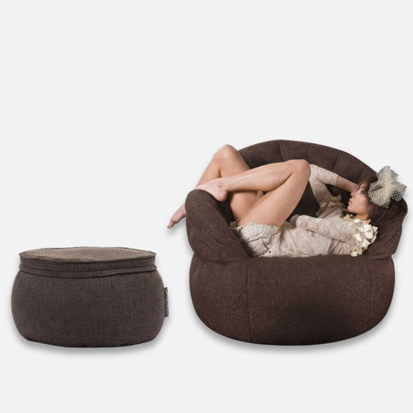 En kvinde i let tøj og hat slapper af i en stor, rund Butterfly Chaise-lænestol. Ved siden af hende står en matchende ottoman i memory foam, begge med et blødt, plysagtigt udseende. Baggrunden er helt hvid.