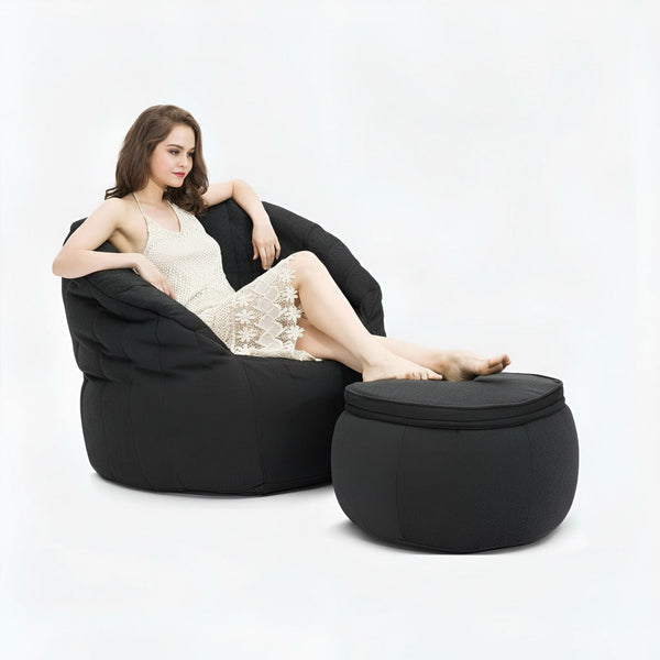 En kvinde i en hvid blondekjole sidder komfortabelt på en sækkestol i memory foam med fødderne hvilende på en matchende sort ottoman på en almindelig hvid baggrund.