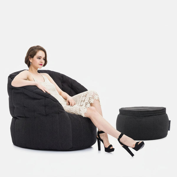En kvinde i en hvid blondekjole og sorte højhælede sko sidder på en stor, mørk Memory Foam Butterfly-sofa med benene over kors. En matchende rund ottoman er placeret ved siden af hende, alt sammen på en almindelig hvid baggrund.