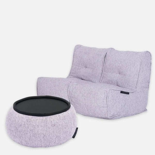 En moderne, lysegrå tvillingesofa i stof med tuftede puder er sat sammen med en matchende rund ottoman med en sort bakkeplade, begge på en ensfarvet lysegrå baggrund.