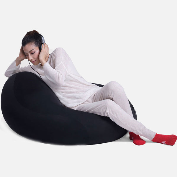 En person i lyst loungewear og røde sokker slapper af på en sort Ambient Lounge-sækkestol og lytter til musik med hovedtelefoner på en almindelig hvid baggrund.