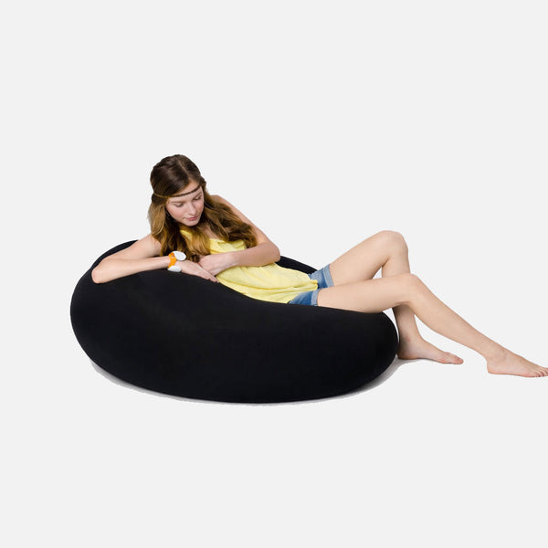 En ung kvinde i gul top og denimshorts slapper af barfodet på en Ambient Lounge Space Pod Sittesekk, en stor, sort, oval sækkekork på en almindelig hvid baggrund.