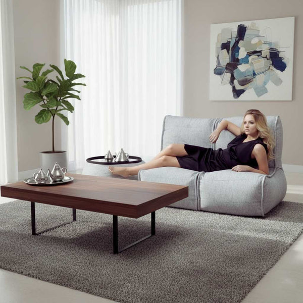 En kvinde i en sort kjole slapper af i en fleksibel sofa i en moderne stue med et sofabord i træ, potteplanter, abstrakt vægkunst og blødt naturligt lys fra høje vinduer.