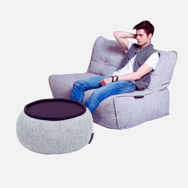 En ung mand sidder afslappet på en moderne, lysegrå fleksibel sofasækkestol med en arm hvilende på ryggen. Foran ham står en matchende rund ottoman med en sort bakke, begge på en almindelig hvid baggrund.
