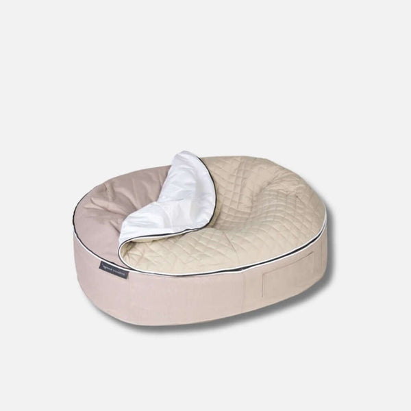 En beige, oval allergivennlig hundeseng med quiltet topptrekk og et hvidt, delvist foldet tæppe på den ene side, placeret på en lysegrå baggrund.