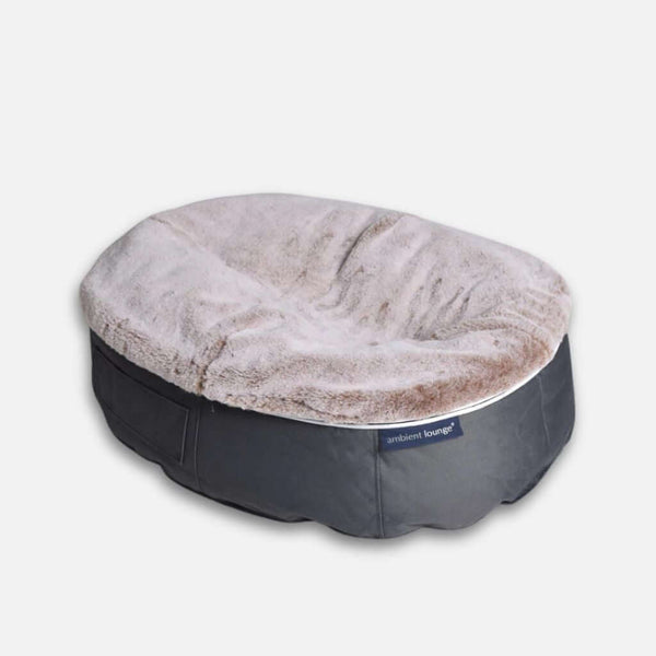 En blød, oval hundeseng med en plysagtig, lys beige pude og en mørkegrå stofbase med et lille Ambient Lounge-mærke på siden for ultimativ komfort for hund.