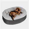 En lille brun hund ligger på en grå, oval hundeseng med quiltet ThermoQuilt-overflade. En blød legetøjsbold ligger ved siden af hunden. Baggrunden er helt hvid.