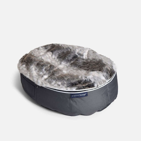 En rund hundeseng med en top af imiteret pels i grå nuancer og en bund af mørkegråt stof, med en etiket, hvor der står hundeseng Ambient Lounge, på en almindelig hvid baggrund.