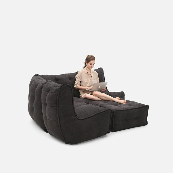 En kvinde sidder barfodet på en stor, mørkegrå designer-hjørnesofa med tilhørende chaiselong, mens hun bruger en bærbar computer på skødet mod en almindelig hvid baggrund.