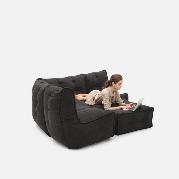 En kvinde ligger på maven i en moderne, mørkegrå designersofa med bøjede ben og bruger en bærbar computer på en skammel mod en almindelig hvid baggrund.