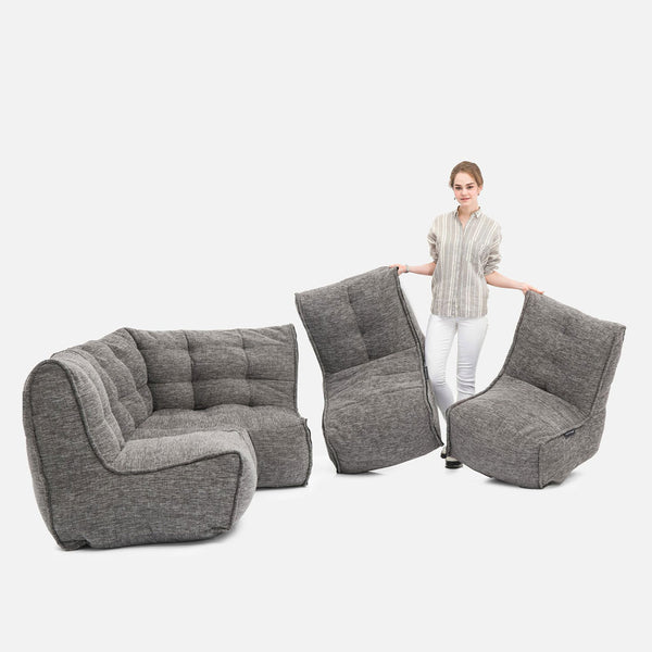 En kvinde står ved siden af en modulær grå Ambient Lounge Mod 4 L-sofa og holder to separate polstrede stole, mens en anden stol og et hjørnestykke er anbragt i nærheden for at vise fleksible møbler på en hvid baggrund.