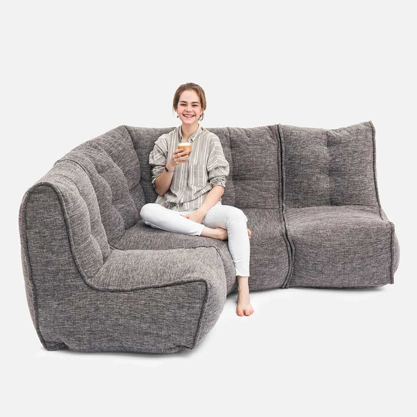 En person sidder barfodet på en grå Ambient Lounge Mod 4 L-sofa, smiler og holder en smartphone mod en almindelig hvid baggrund - og viser, hvordan fleksible møbler kan forbedre ethvert moderne opholdsrum.