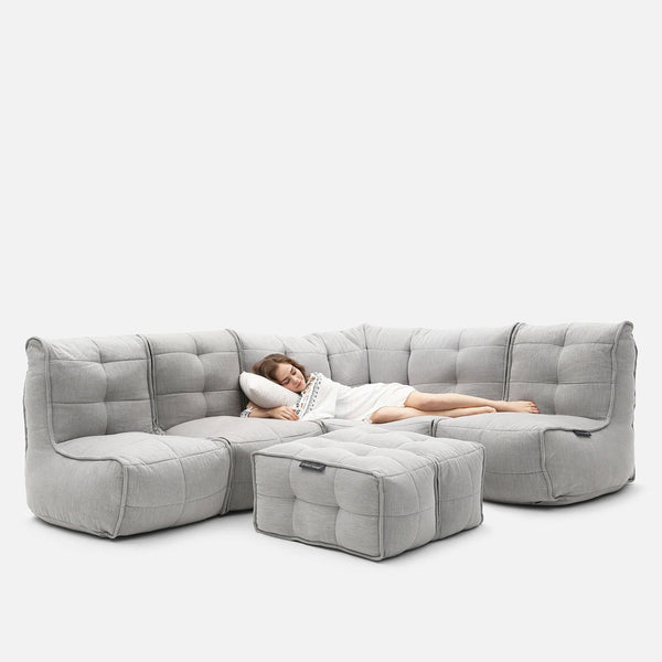 En kvinde i en hvid kjole ligger på en lysegrå, tuftet Modulsofa med matchende ottoman og hviler hovedet på en pude i en afslappet stilling. Det alsidige design blander komfort og stil, perfekt til moderne rum. Baggrunden er almindelig hvid.