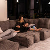 Et par slapper af i en stor grå Ambient Lounge modulær sofa om aftenen. Manden ligger med hovedet i kvindens skød og smiler, mens hun forsigtigt lægger sin hånd på hans bryst. Byens lys skinner ind gennem vinduerne i denne moderne stueløsning.