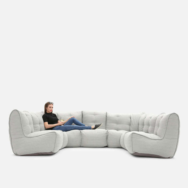En person sidder afslappet med strakte ben i en stor, moderne Ambient Lounge-sædesofa med tuftede puder på en almindelig hvid baggrund.