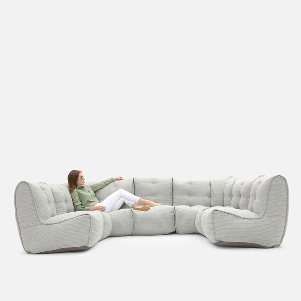 En kvinde i grøn top og hvide bukser sidder afslappet i en stor, lysegrå Ambient Lounge U-formet sofa mod en almindelig hvid baggrund.