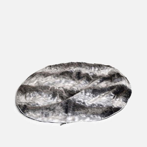 En rund, plysset hundeseng med en luksuriøs pelstopp og fuskepelstrekk i blandede nuancer af grå, sort og hvid, placeret på en ensfarvet lys baggrund.