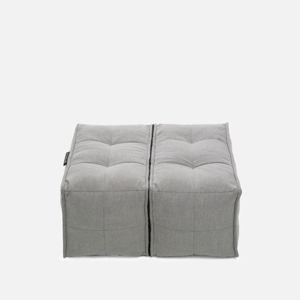To firkantede, grå, tuftede ottomaner fra Ambient Lounge er placeret side om side på en almindelig hvid baggrund og viser deres designede ottoman-stil og slanke, synlige sømme.