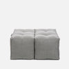 To grå, firkantede Twin Ottomans fra Ambient Lounge har tuftede detaljer og synlige sømme, arrangeret side om side på en almindelig hvid baggrund for et moderne designer-ottoman-look.