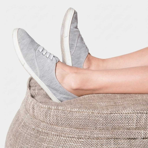 En person med bare ben hviler fødderne iført lysegrå lærredssneakers med hvide såler på en struktureret beige Wing Ottoman mod en almindelig lys baggrund.