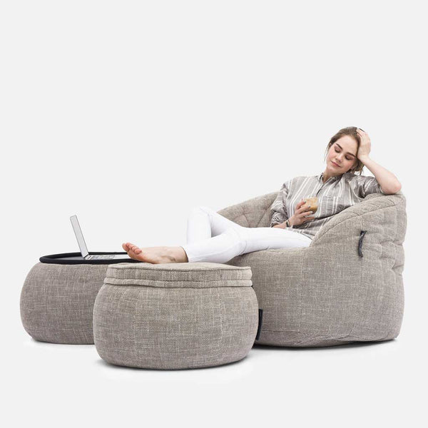 En kvinde slapper af i en lysegrå Butterfly-sofa med fødderne oppe på en matchende ottoman, mens hun holder en drink i den ene hånd. En bærbar computer ligger åben på en anden ottoman. Baggrunden er helt hvid.