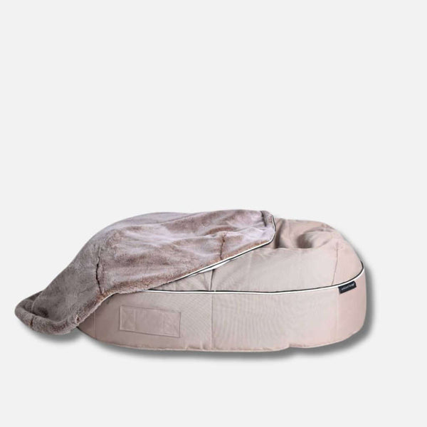 En lys beige, oval kjæledyrseng med hævede sider, delvist dækket af et blødt, gråbrunt fuskepelstrekk, på en ensfarvet hvid baggrund.