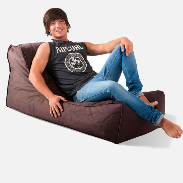 En ung mand med brunt hår, iført en sort ærmeløs Rip Curl-skjorte og blå jeans, smiler og læner sig tilbage på en brun Studio Lounger-sæk mod en almindelig hvid baggrund.